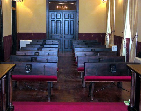 Sala con bancos de madera y tapicería roja, puertas al fondo.